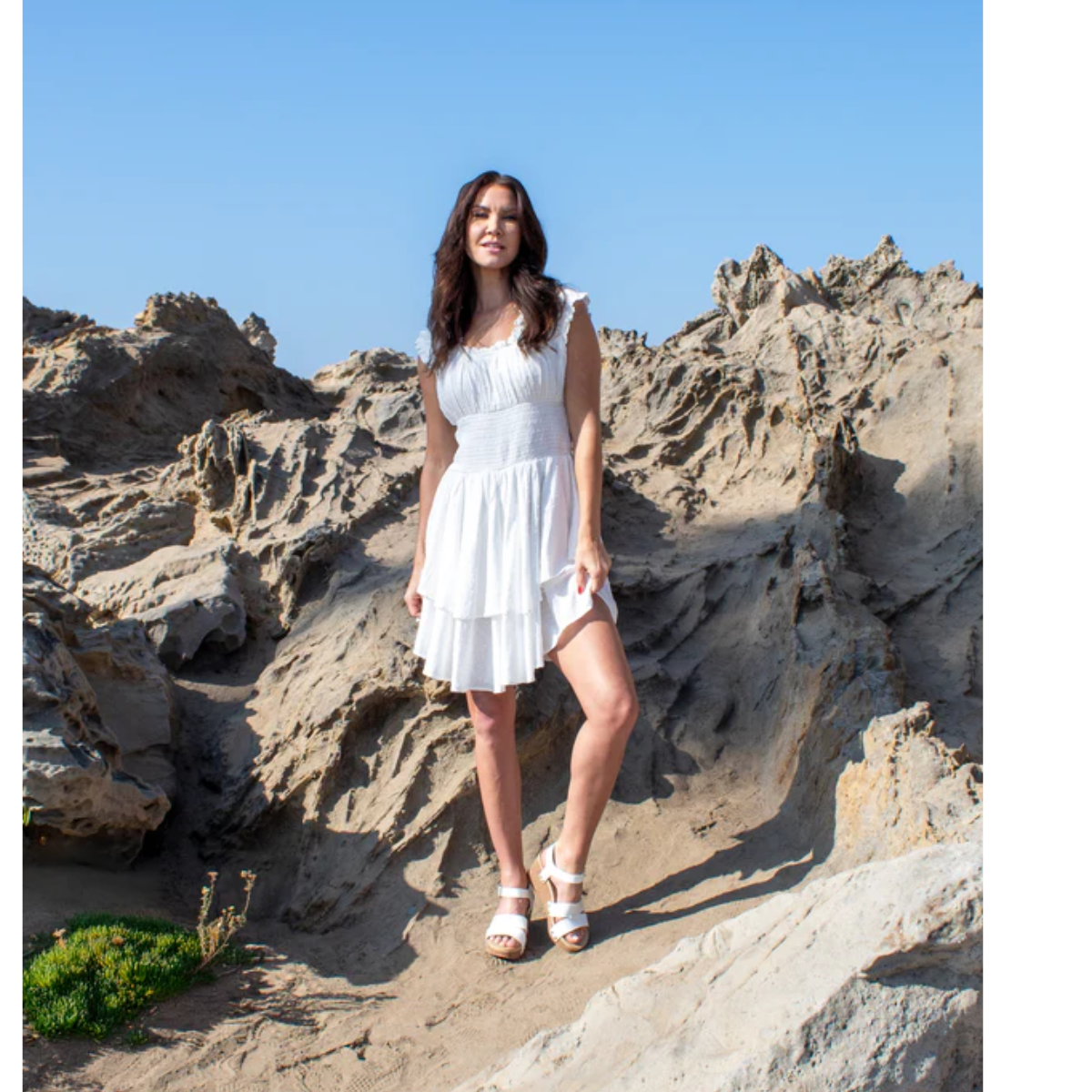 A woman in a white dress confidently stands on the rocky, sunlit terrain under a clear blue sky, elevated by Volatile's Agnes Wedges Sandal in White.