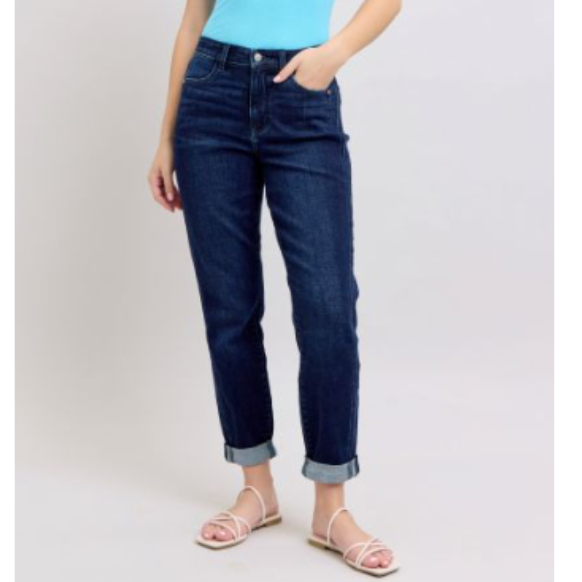 A person in cuffed Judy Blue High Waisted Boyfriend Jeans and a turquoise top stands with one hand in their pocket. White sandals complete the relaxed look against a plain background.