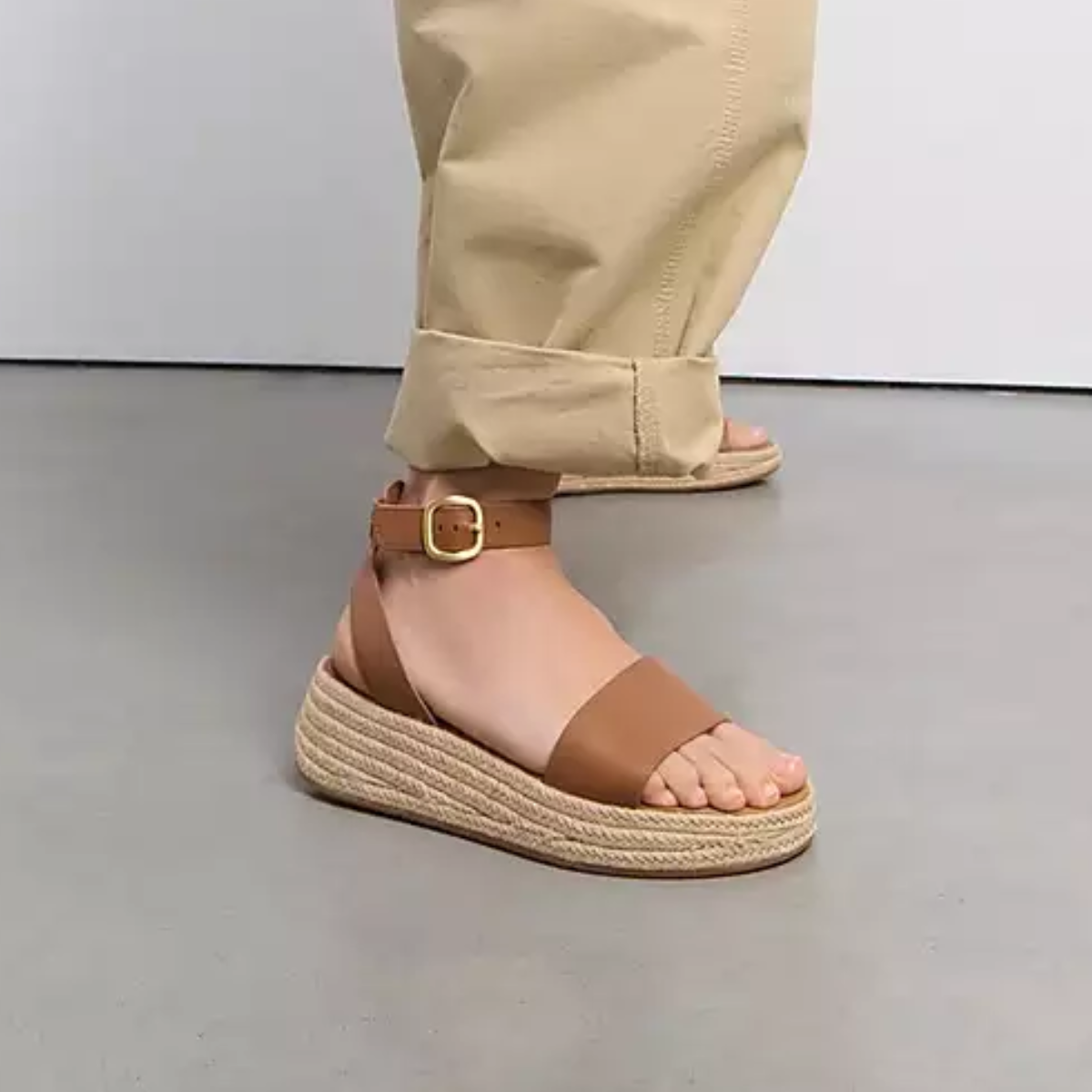 A person wearing beige cuffed pants and FITFLOP USA LLC's F-MODE Espadrille Leather Flatform Ankle-Strap Sandals with a Microwobbleboard midsole stands on a gray floor.
