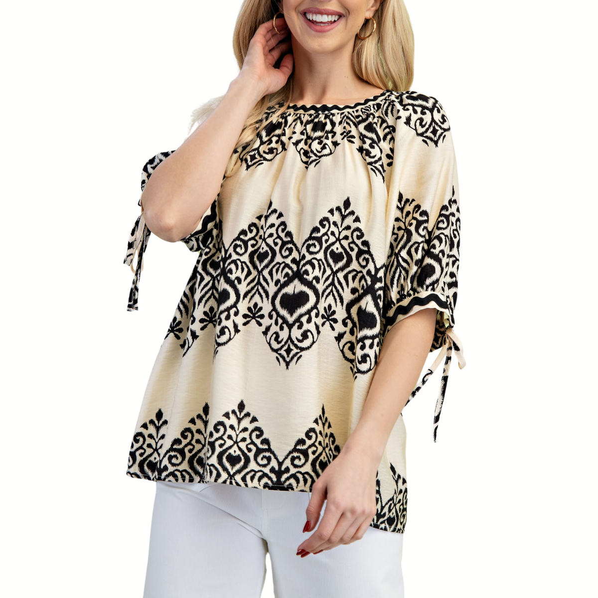 A woman smiles, touching her hair, wearing the Fahiongo Round Neck Short Sleeve Top with Ric Rac Trim in a black and beige pattern, paired with white pants.