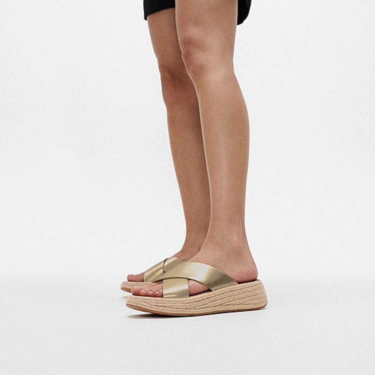 A person stands against a plain background wearing black shorts and FITFLOP USA LLC's F-MODE Espadrille Metallic-Leather Flatform Cross Slides, featuring a braided platform sole and gold crisscross strap, adding a chic touch to their stylish ensemble.