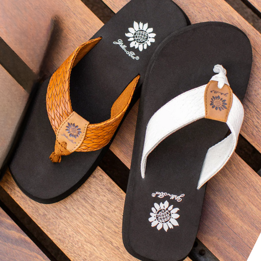 A pair of Flax Flip Flop sandals by Yellow Box on a wooden surface, one with a brown faux leather strap and the other with a white strap, both featuring the signature sunflower logo on the insole.