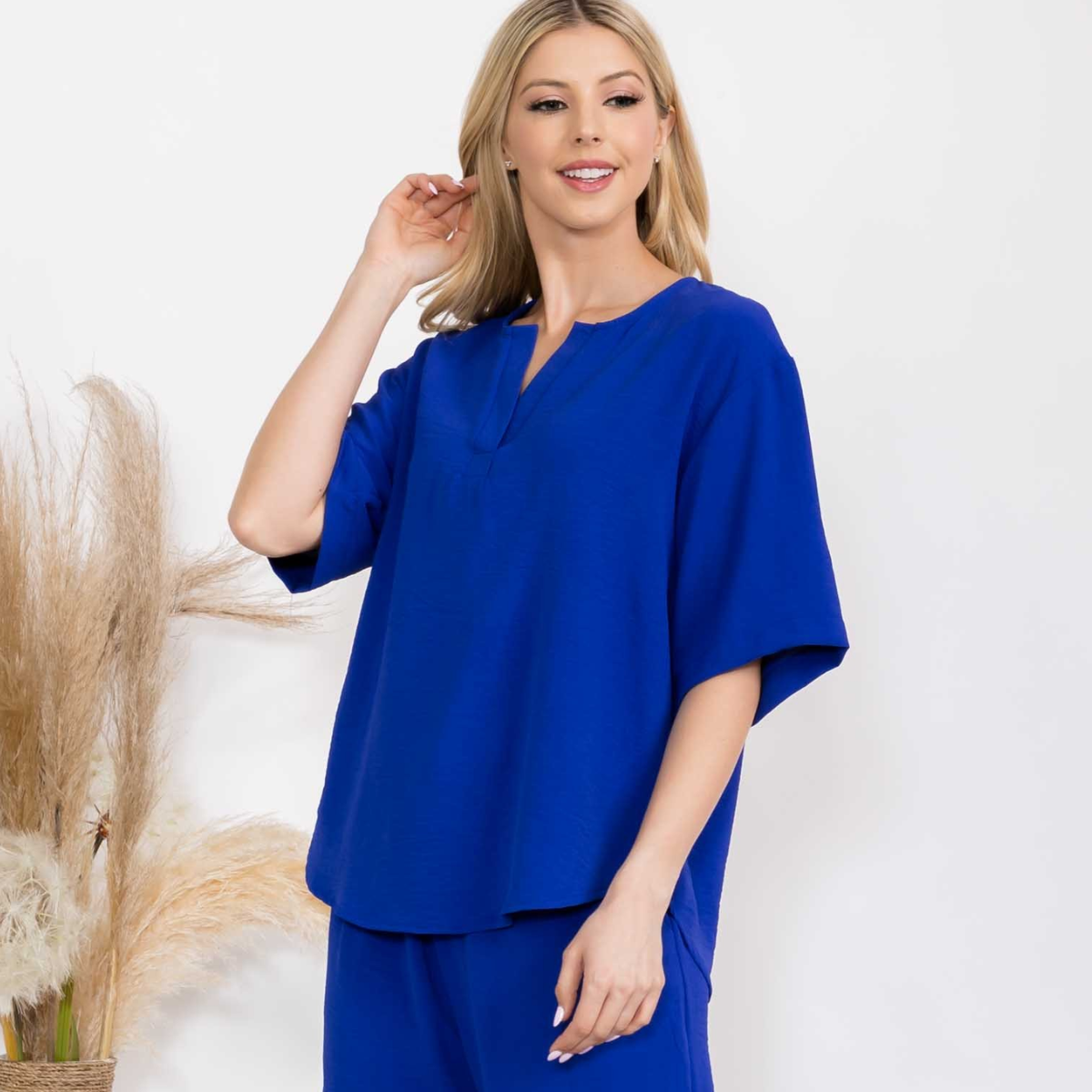 A woman poses indoors, smiling with one hand by her hair, wearing the FASHION GO Split Neckline Air Flow Top in Royal Blue and matching pants. Pampas grass decor accents the background.