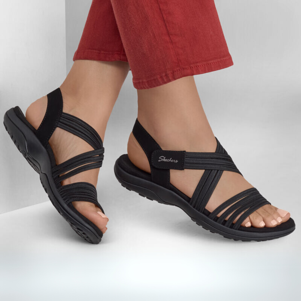 Against a white background, a person models the SKECHERS Reggae Slim Coffee Run in Black. These black strappy sandals have a slight wedge heel and pair elegantly with red pants. Made from vegan materials, they offer an ethical choice and ensure all-day comfort.