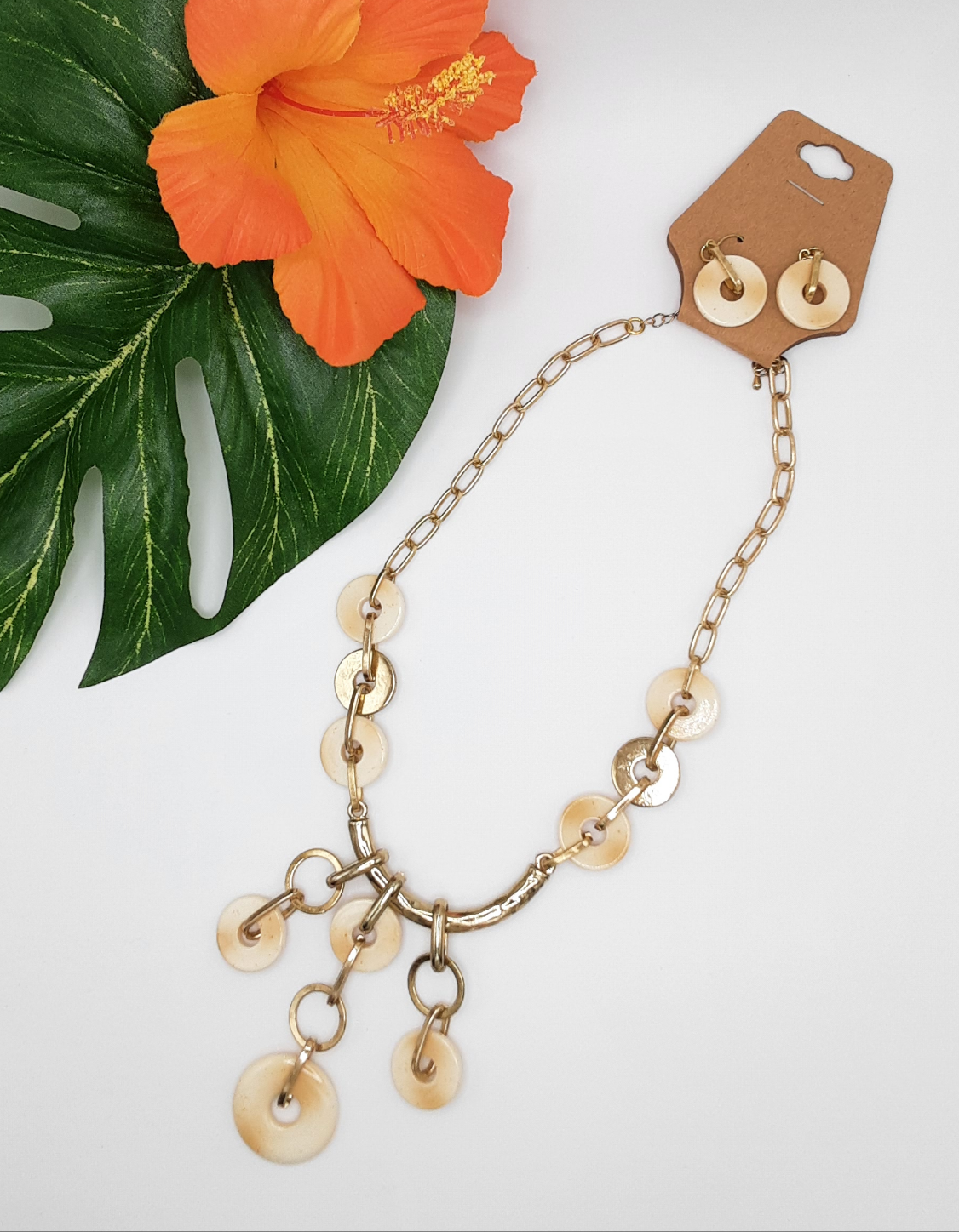The Beige "donuts" and Gold Chain Link 17 inch Necklace Set by SPECIAL EFFECTS features a gold chain necklace with circular pendants, paired with matching earrings, all displayed on a white background alongside a green leaf and an orange flower.