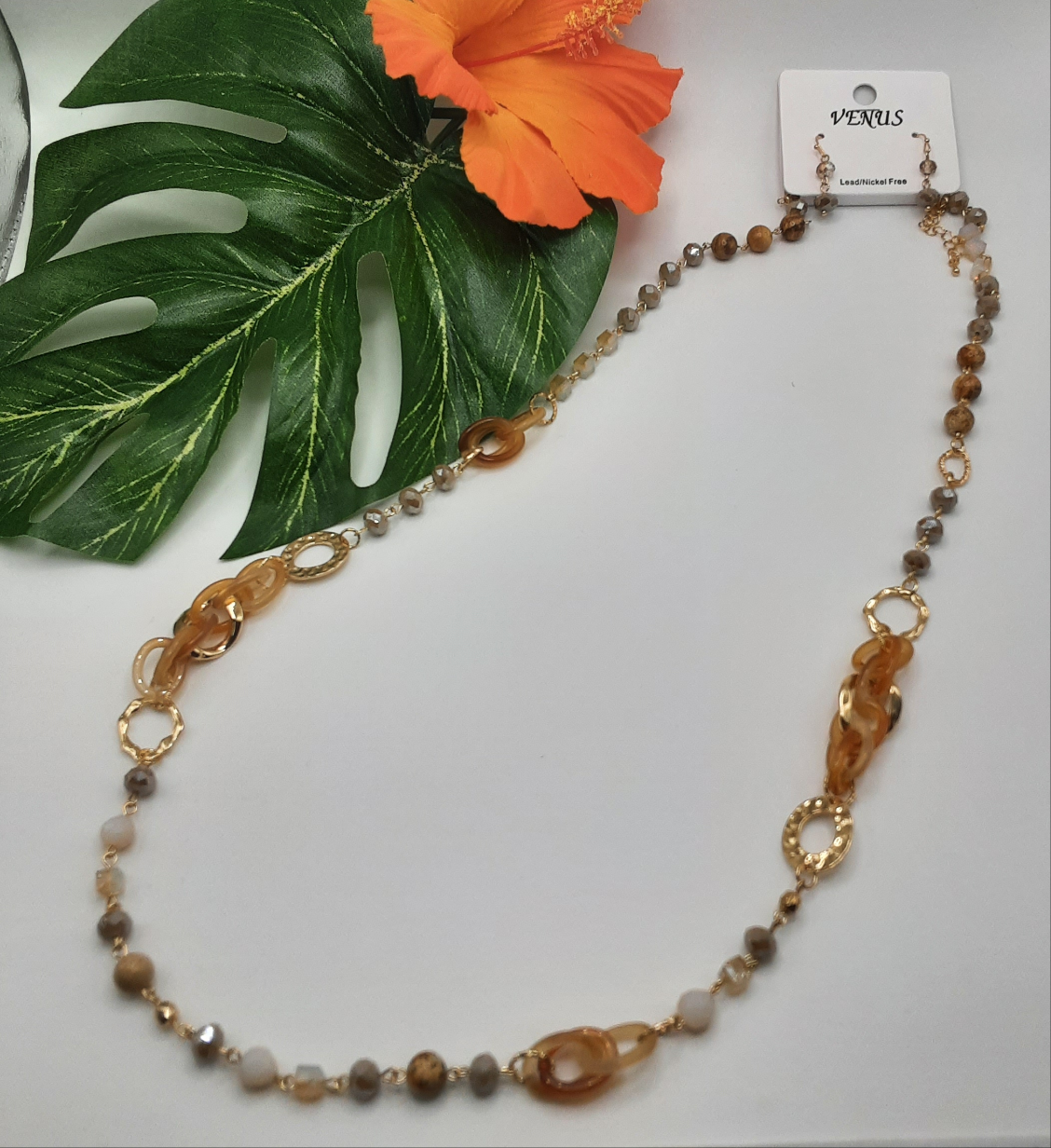 A 36-inch necklace from SPECIAL EFFECTS, featuring Topaz Beads and Gold Oval Chain Links, is elegantly displayed on a white surface, accompanied by a large green leaf and an orange hibiscus flower. A label reads "Venus".