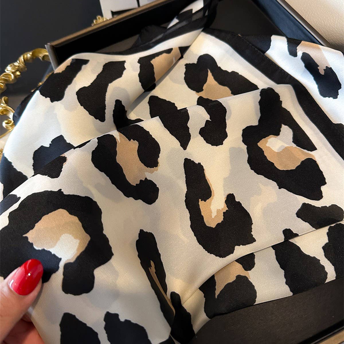 A hand holds the ACCITY Leopard Print Satin Square Scarf, a chic black, beige, and brown accessory perfect as a hair wrap or stylish addition to any outfit.