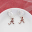Seasons Jewelry’s Alabama Glitter Enamel Logo Huggie Hoop Earrings—silver hoops adorned with red and silver letter "A" charms and sparkling crimson glitter enamel—are showcased on a white plate against a textured red and white backdrop.