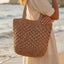 A person in a light-colored dress stands on the beach, holding the Anarchy Street Large Woven Straw Fan Stitch Shoulder Tote Bag.