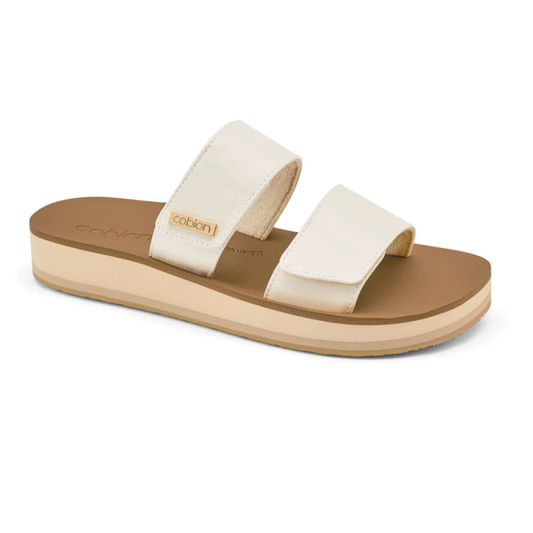 A pair of Dana Rise slide sandals in white by Cobian with tan soles, featuring adjustable straps over a flat base.