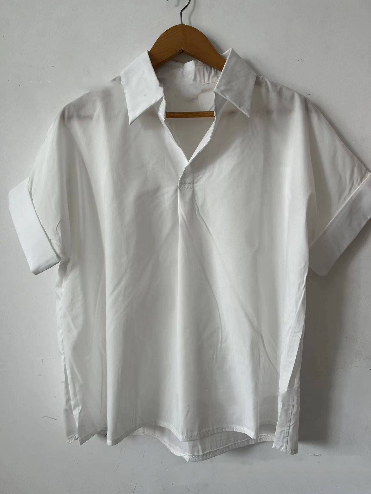 The Rosa Clothing White Relaxed Short Sleeve Blouse, featuring a chic collar, is displayed on a wooden hanger against a plain white wall for an effortlessly stylish look.