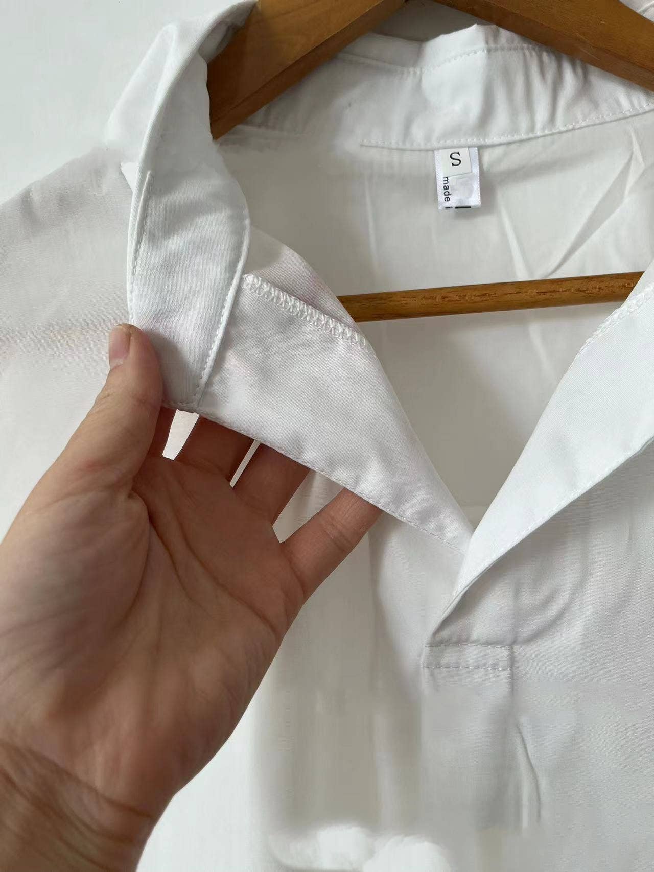 A hand holds the collar of a Rosa Clothing White Relaxed Short Sleeve Blouse on a wooden hanger, showing the size small tag inside the neckline.