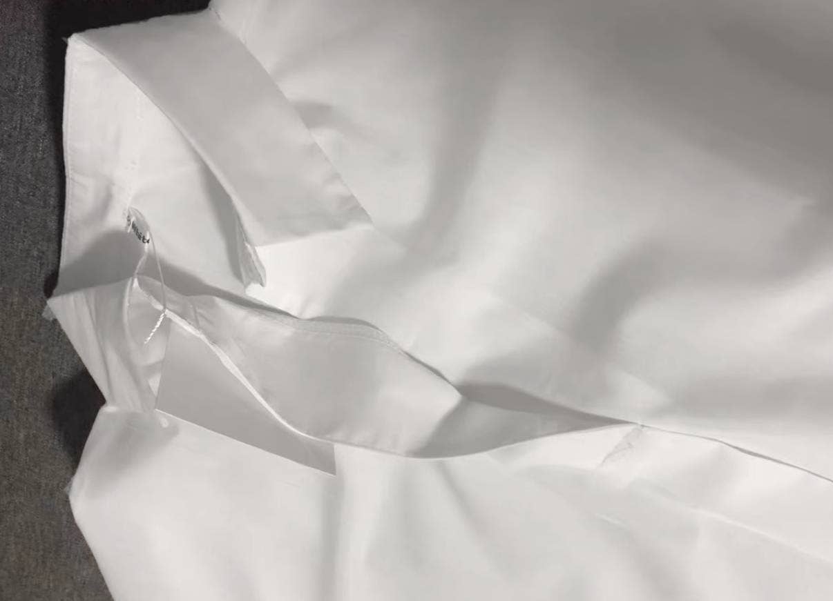 A close-up of the Rosa Clothing White Relaxed Short Sleeve Blouse with a visible tag, laid flat on a gray surface.