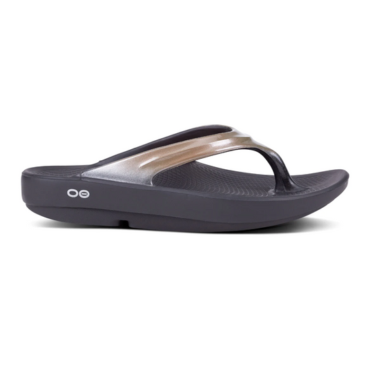 A single black OOFOS LLC OOLala Lux in Latte flip-flop with a metallic strap and strategic arch support is isolated on a white background.