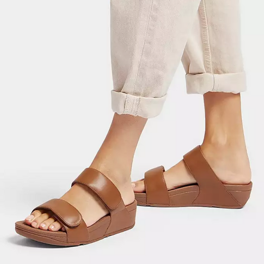 A person showcases rolled-up beige pants and minimalist-chic Lulu Adjustable Leather Slide Sandals in Light Tan by Fit Flops, featuring thick straps and possibly Microwobbleboard™ midsoles for added comfort, all set against a white background.