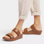 A person showcases rolled-up beige pants and minimalist-chic Lulu Adjustable Leather Slide Sandals in Light Tan by Fit Flops, featuring thick straps and possibly Microwobbleboard™ midsoles for added comfort, all set against a white background.