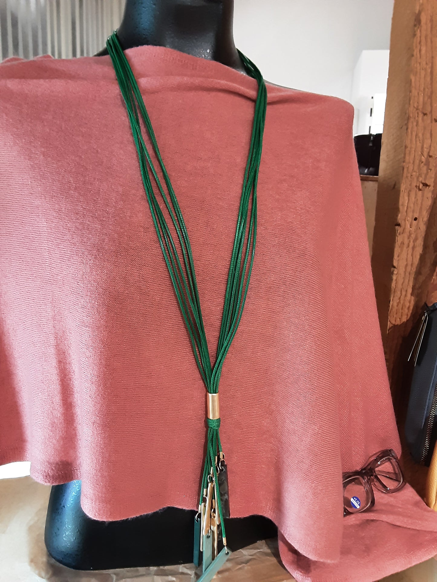 A mannequin draped in a pink fabric displays the Multi Row Green Tassel Long Necklace Set by SPECIAL EFFECTS, featuring multiple green strands with a gold accent, elegantly hanging down.