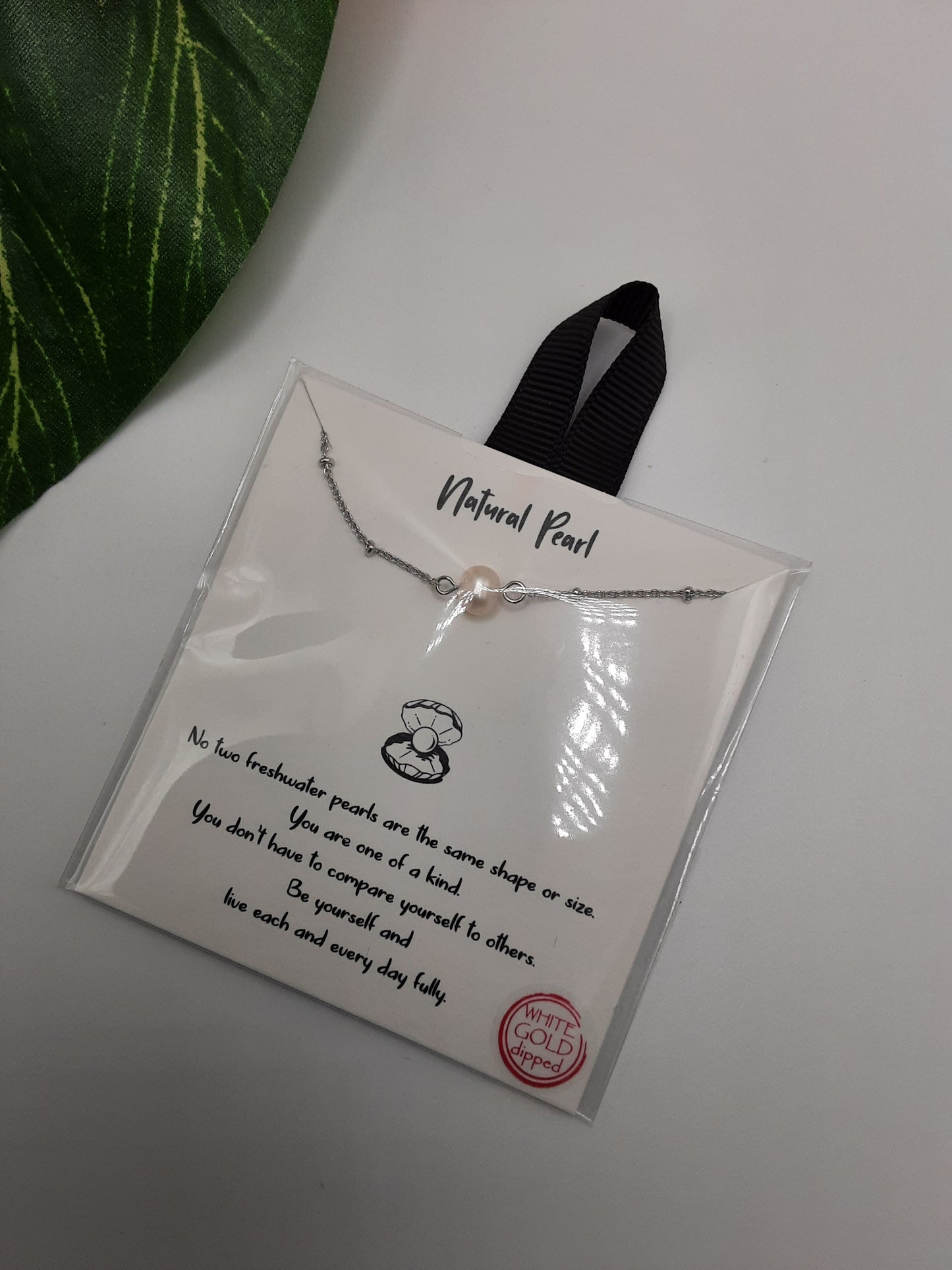 A Natural Pearl 16 inch Carded Necklace by SPECIAL EFFECTS, featuring a pearl pendant on a silver chain, lies on a white surface near a green leaf. The package text emphasizes the uniqueness of freshwater pearls with an inspirational message.