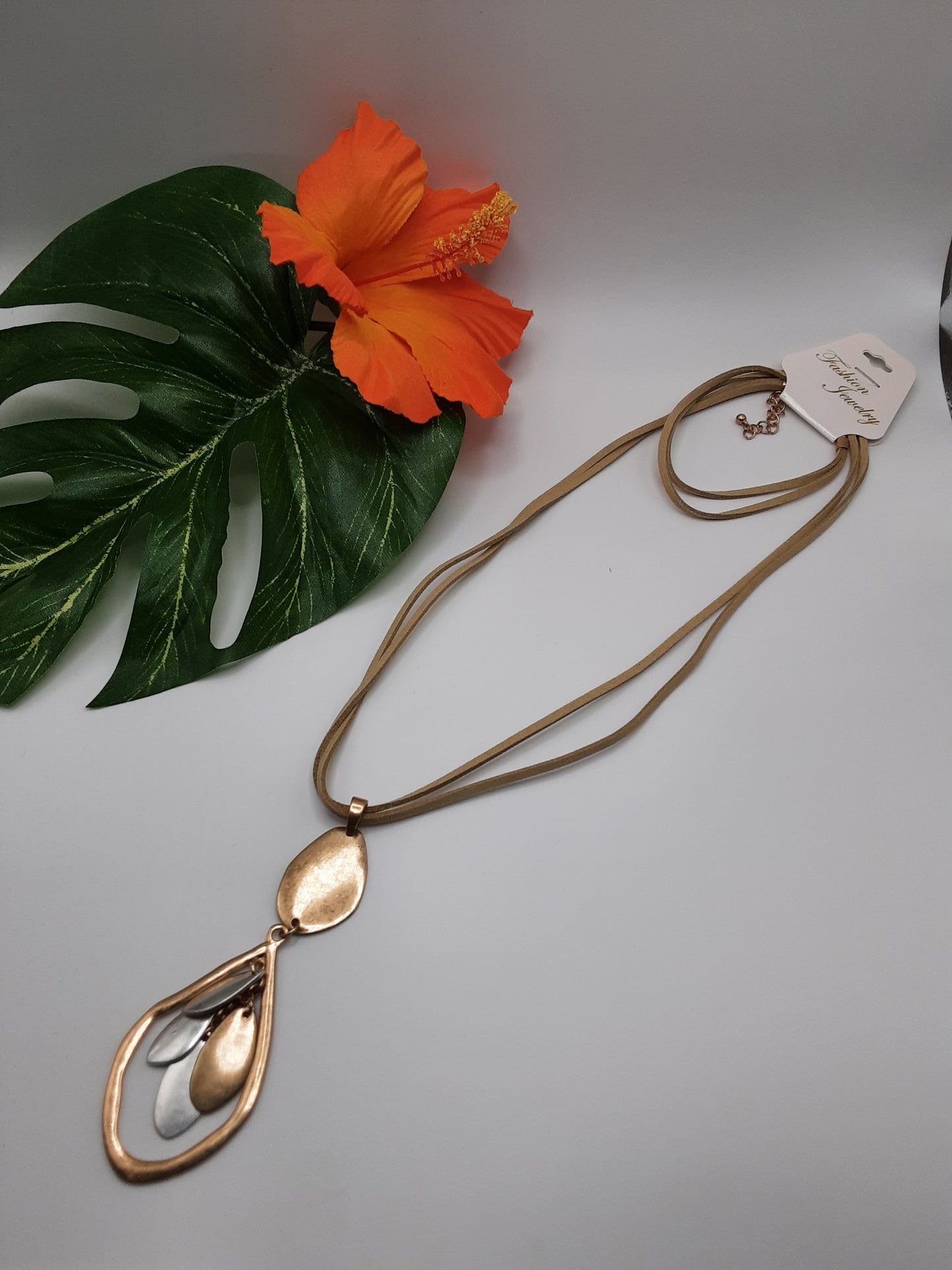 A 2 Tone Metal Tear Drop Necklace with Tan Cord by SPECIAL EFFECTS lies next to a green Monstera leaf and a vibrant orange hibiscus flower.
