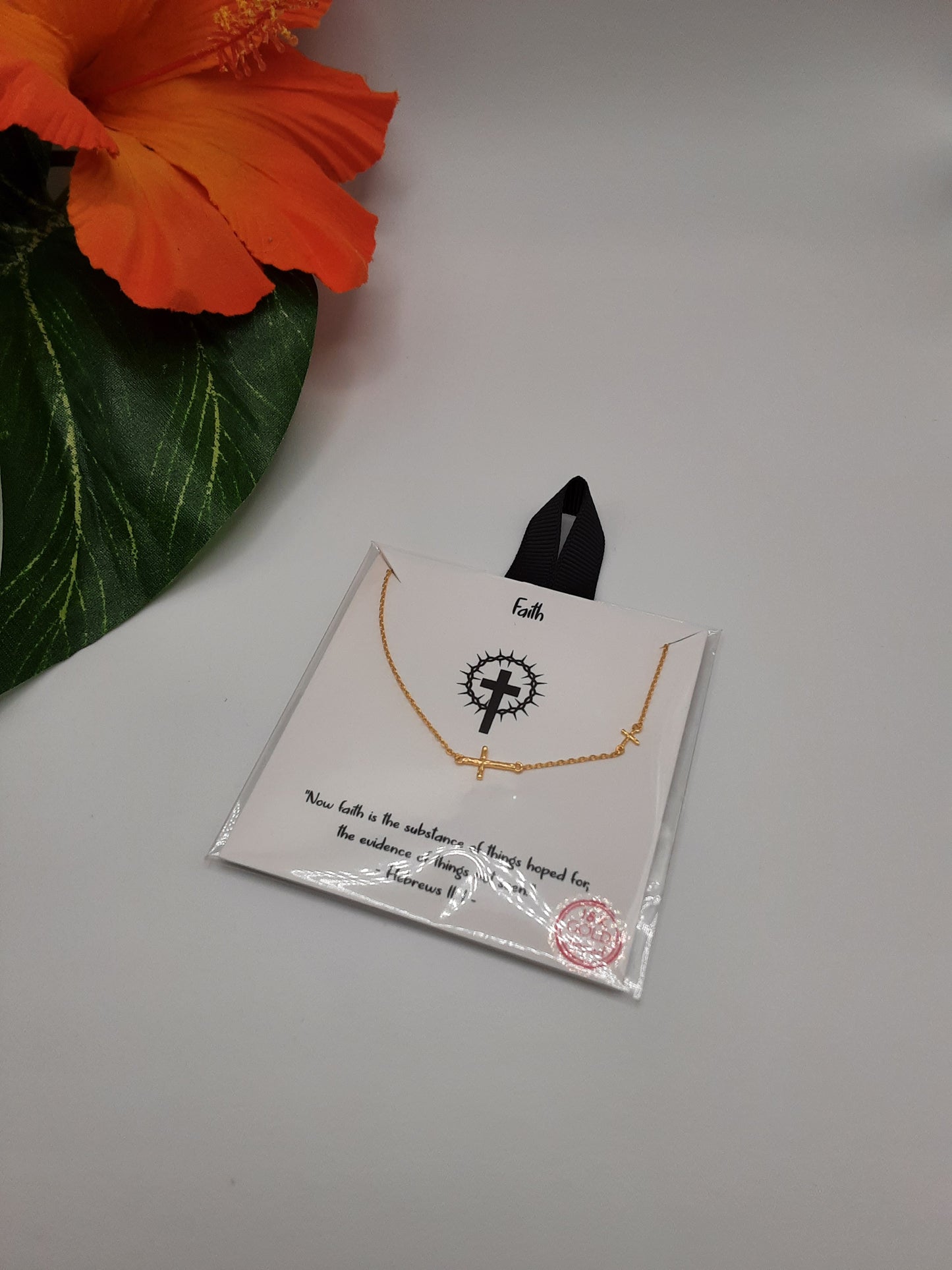 The Faith Small Crosses on Chain Necklace Gold dipped by SPECIAL EFFECTS is displayed on a white card, adorned with a black ribbon and script text. Nearby, an orange flower with green leaves adds a touch of color.