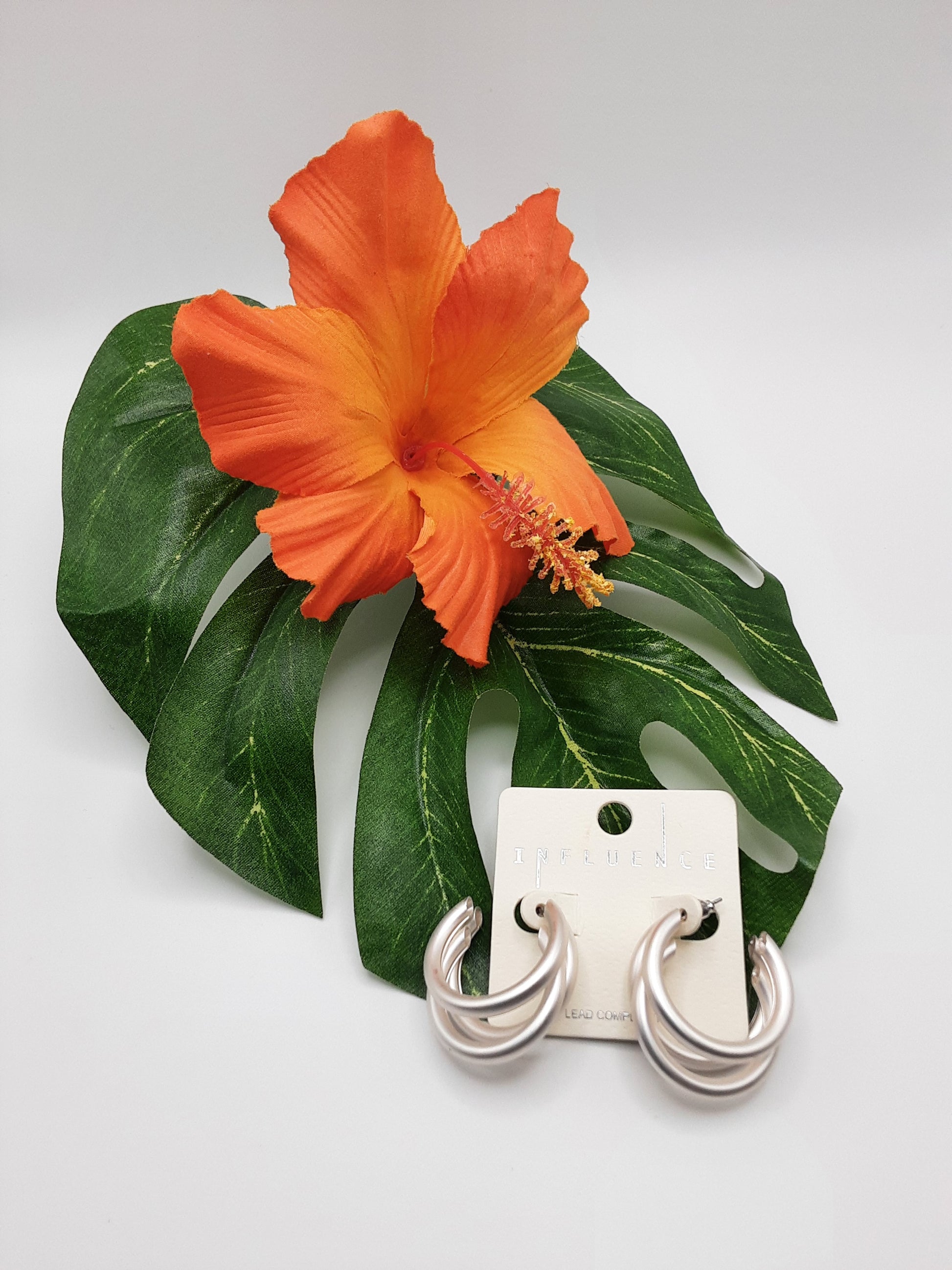 Matte Metal Tube Hoop Earrings by SPECIAL EFFECTS elegantly sit on a white card next to a green leaf and an orange hibiscus flower.