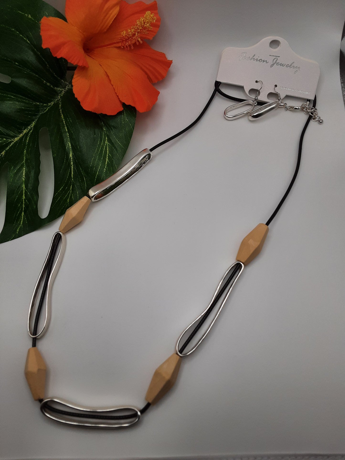 The 5 Station Natural Wood Large Bead and Black Cord Long Necklace Set by SPECIAL EFFECTS is displayed on a white surface, complemented by an orange hibiscus flower and green leaf in the background.