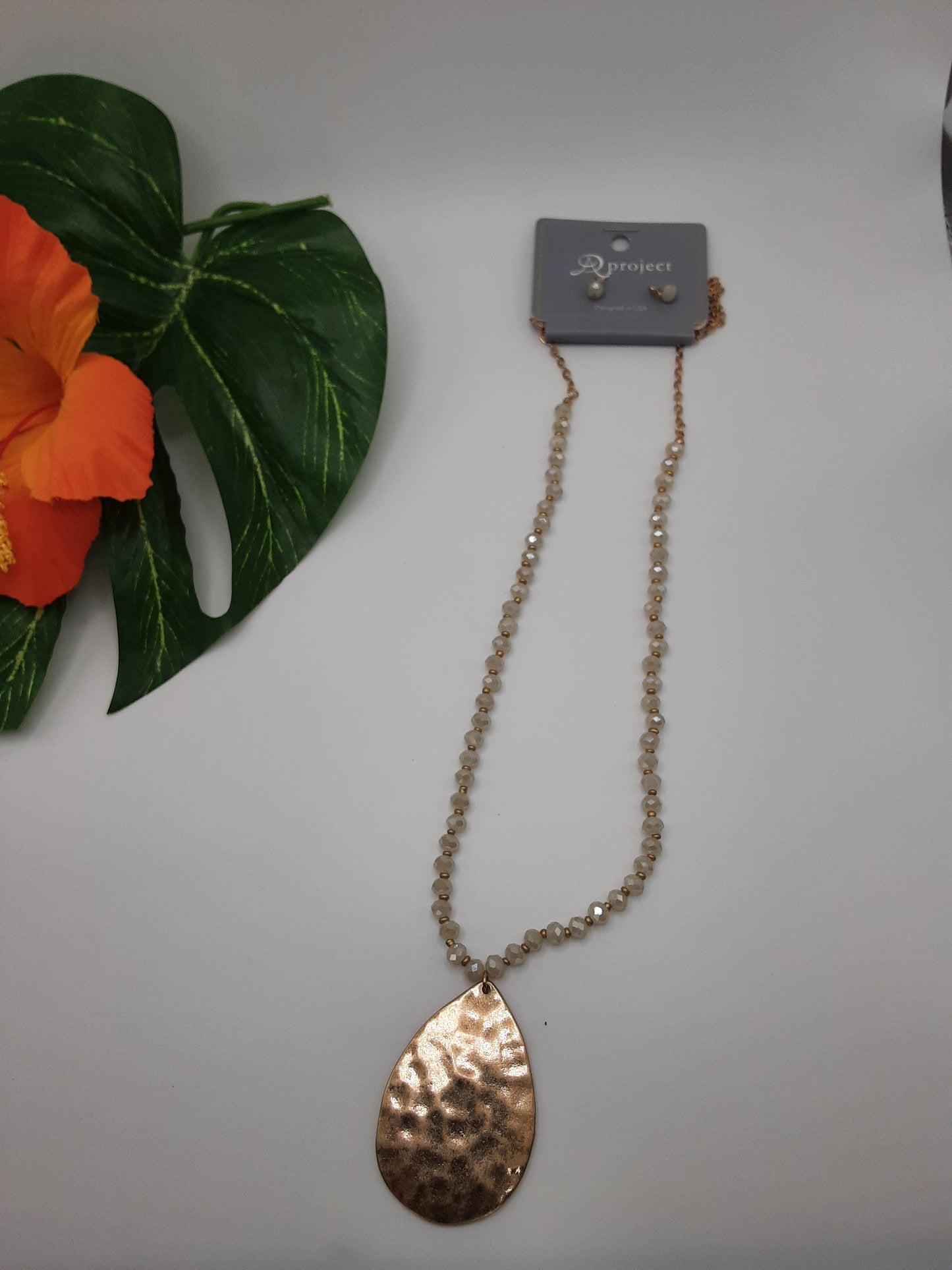 The SPECIAL EFFECTS Teardrop Pendant and Crystal Necklace, featuring a variety of bead colors and a prominent textured teardrop pendant, is shown on a white surface beside an artificial orange flower and a green leaf.