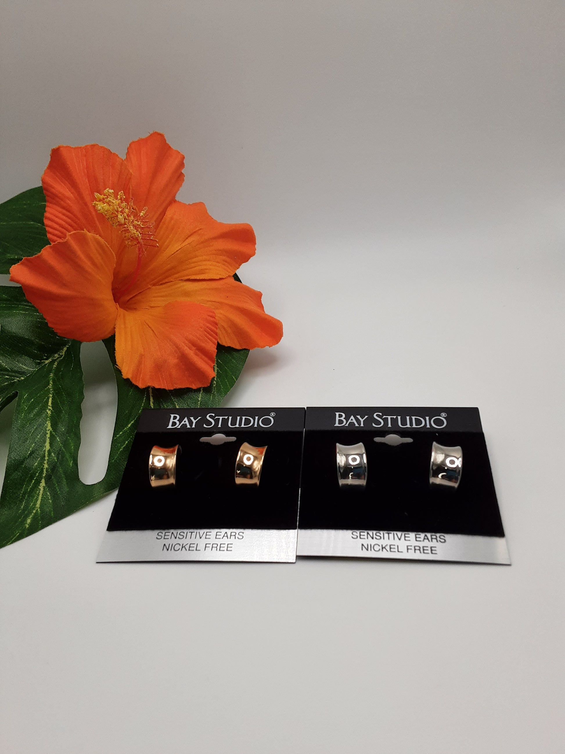 Two pairs of BAY STUDIO 9201EP NICKEL FREE earrings, featuring gold-toned and silver-toned wide hoop designs, nestle next to an orange hibiscus with green leaves. All pieces from SPECIAL EFFECTS are nickel-free for comfort.