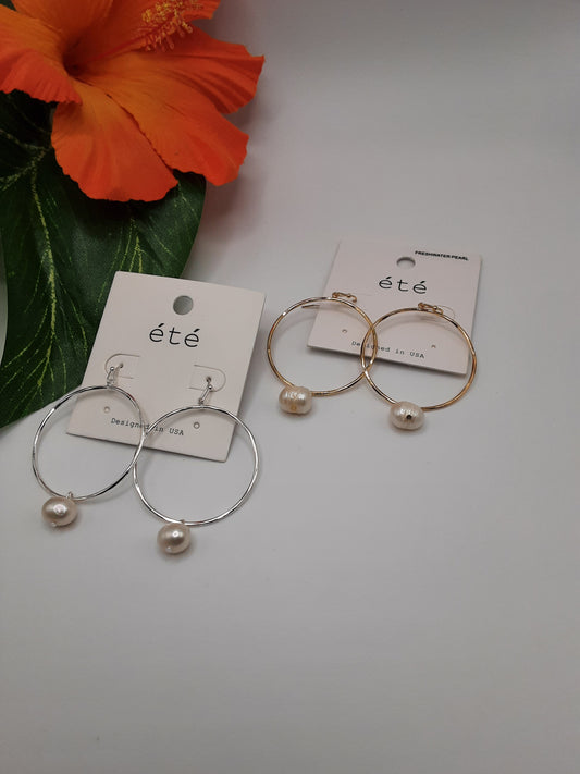 Two pairs of Special Effects' Wire Circle Earrings with Pearl Drop are elegantly showcased on earring cards labeled "été." One pair features a silver finish, while the other boasts a gold hue. An orange hibiscus flower is delicately placed beside the earrings, enhancing their allure.