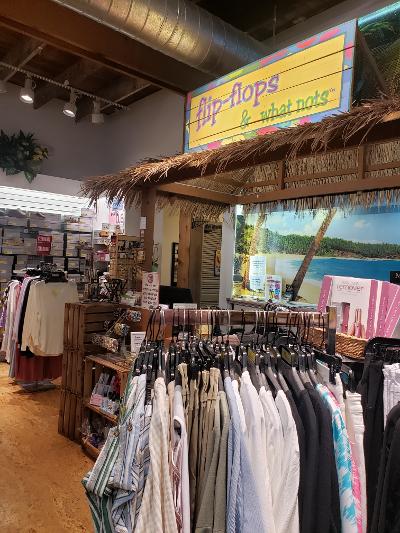 Clothing store interior with racks of shirts and blouses, a tropical-themed sign reading “flip-flops & what nots,” and a beach mural in the background.