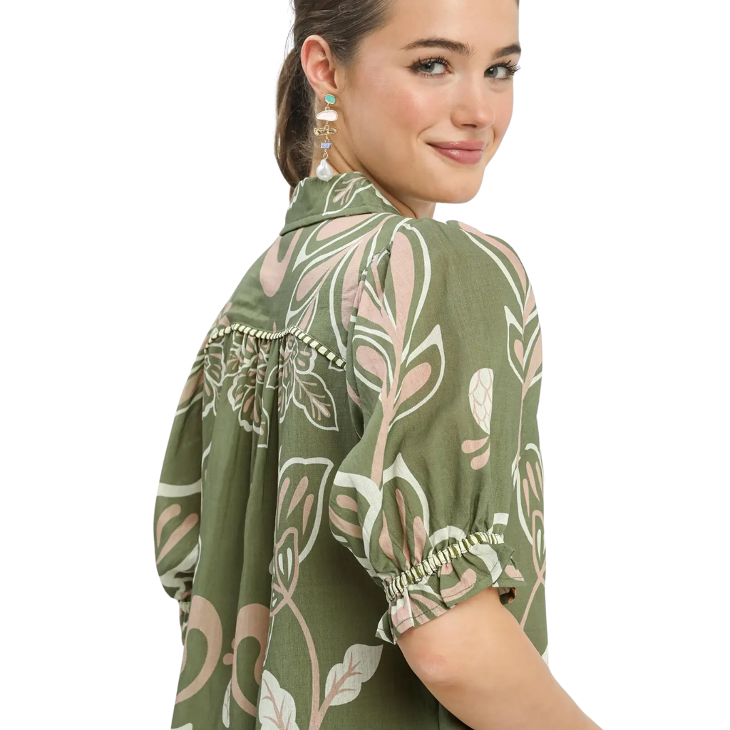 A woman wearing the Umgee Floral Print Puff Sleeve Top in olive green with pearl earrings, glancing over her shoulder against a white background.