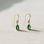 A pair of Ellemadera Chic Gemstone Dangle Earrings featuring green teardrop-shaped gemstones and gold hooks, displayed on a light-colored textured surface.