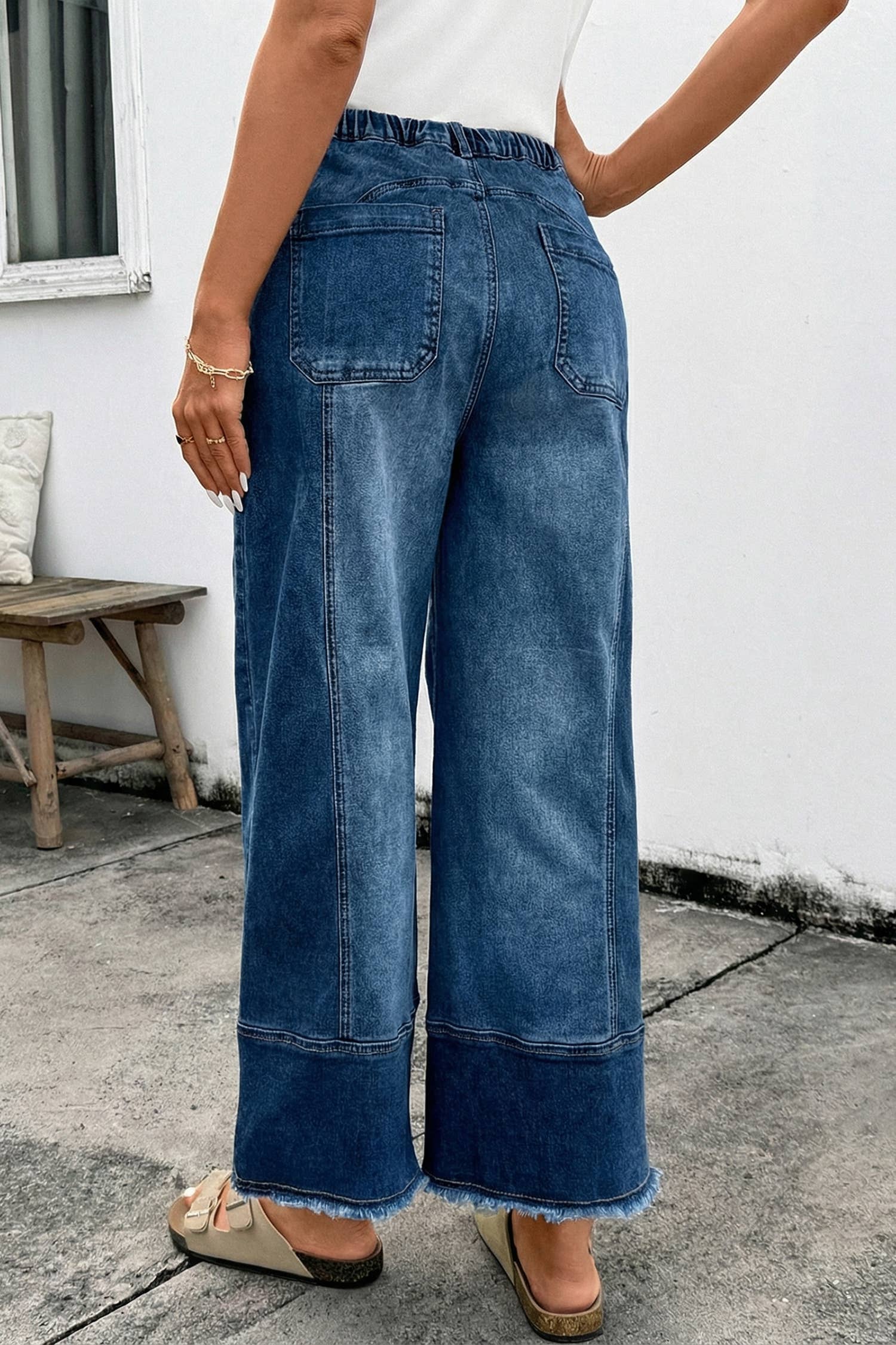 A person wears 1 the Woman Drawstring Seam Detail Raw Hem Wide Leg Denim Pants, beige sandals, and a white top while standing outdoors near a bench and a white wall.