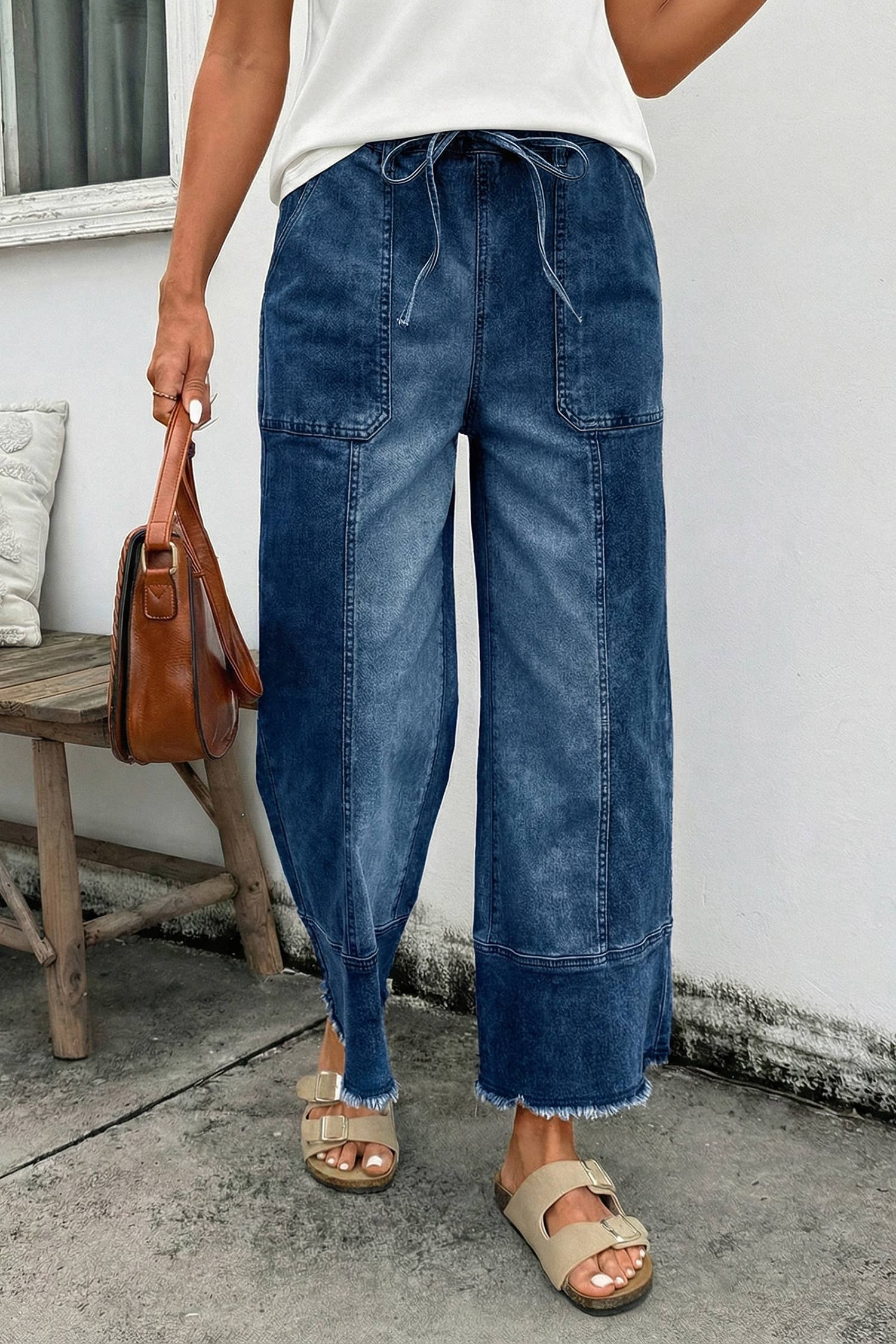 A person in 1 the Woman Drawstring Seam Detail Raw Hem Wide Leg Denim Pants, a white top, beige sandals, and holding a brown handbag stands on concrete near a wooden bench and white wall.