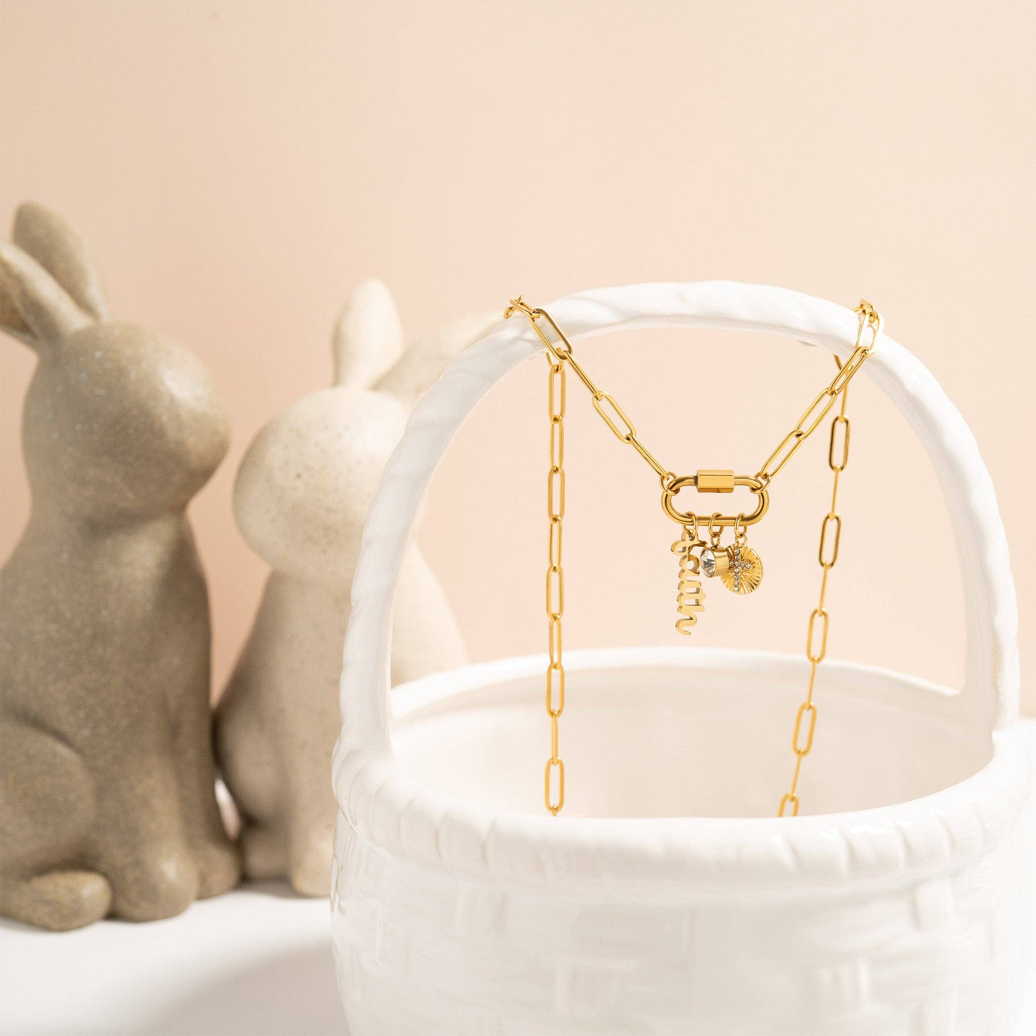The WJW 18K Gold PVD Paperclip Carabiner Necklace is elegantly draped over a white basket, with two decorative stone rabbits in the background against a pale backdrop—perfect as a customizable necklace or Carabiner Charm Holder.
