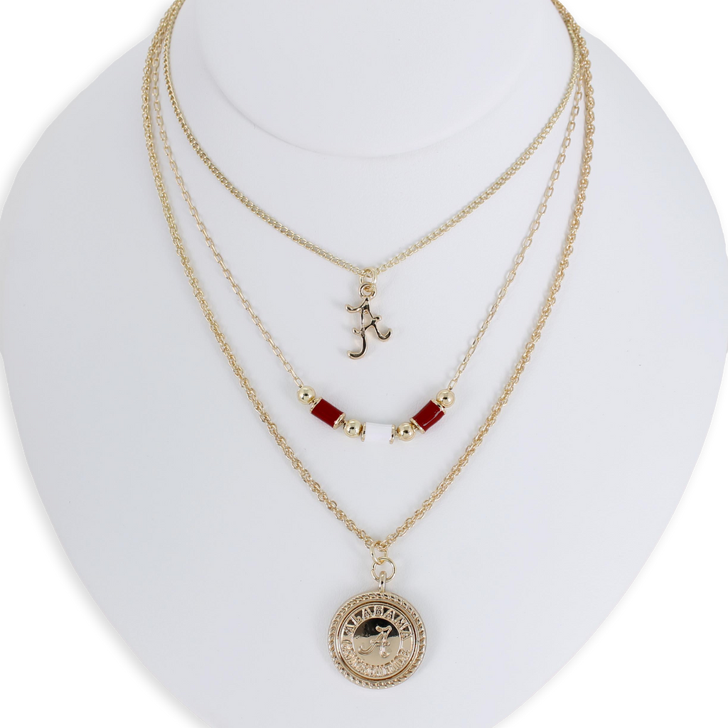 The Alabama Gold & Tube Bead Trio Necklace Set by Seasons Jewelry is showcased on a white bust, featuring three layered gold necklaces: one with an "A" pendant, another adorned with crimson tube beads inspired by Bama jewelry, and a third with a round, detailed charm for a sophisticated collegiate look.