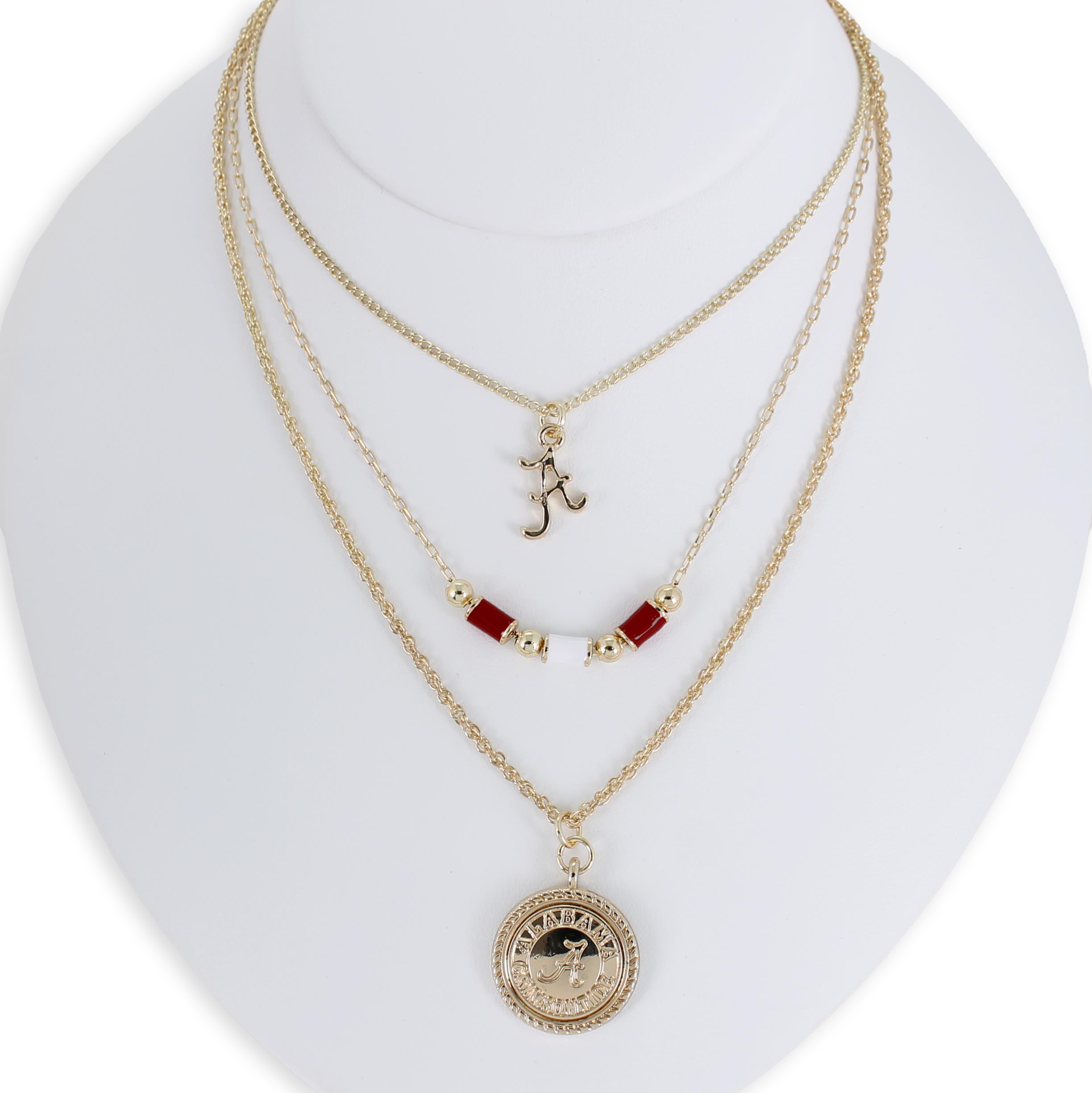 The Alabama Gold & Tube Bead Trio Necklace Set by Seasons Jewelry is showcased on a white bust, featuring three layered gold necklaces: one with an "A" pendant, another adorned with crimson tube beads inspired by Bama jewelry, and a third with a round, detailed charm for a sophisticated collegiate look.