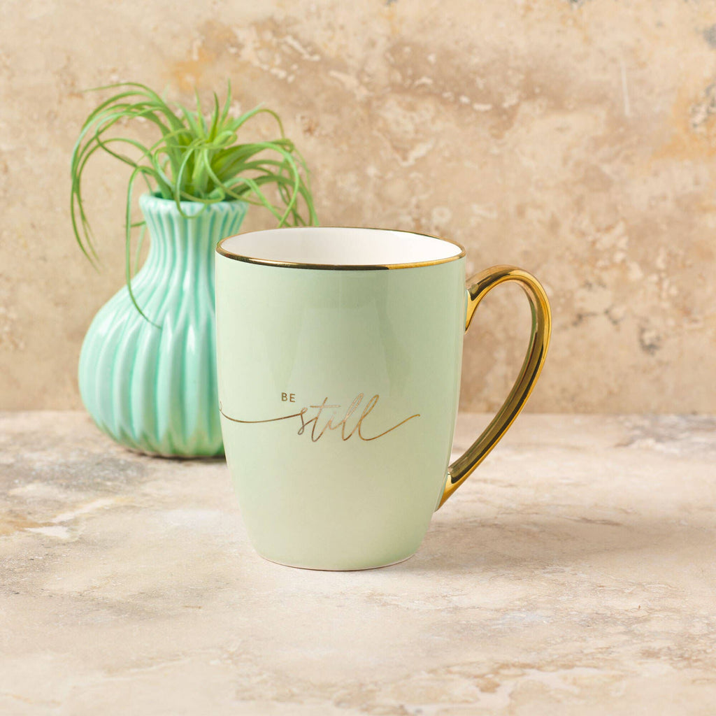 A Mug Mint/Cream Be Still & Know from Christian Art Gifts, featuring a gold handle and inspirational "Be still" text, sits on a stone surface beside a small green vase with a plant—making it the perfect mug to inspire your morning routine.