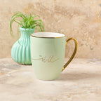 A Mug Mint/Cream Be Still & Know from Christian Art Gifts, featuring a gold handle and inspirational "Be still" text, sits on a stone surface beside a small green vase with a plant—making it the perfect mug to inspire your morning routine.