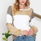 A blonde woman wearing the Shewin Khaki Color Block Exposed Seam Long Sleeve Top and jeans stands in front of a white background. She is smiling slightly with her hands clasped. A potted plant adds a touch of green to the overall khaki color palette in the scene.