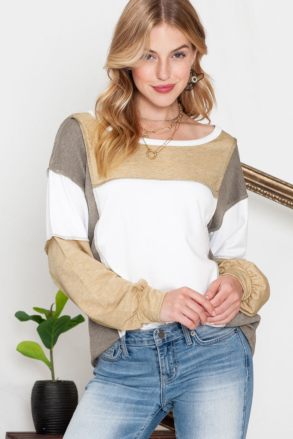 A blonde woman wearing the Shewin Khaki Color Block Exposed Seam Long Sleeve Top and jeans stands in front of a white background. She is smiling slightly with her hands clasped. A potted plant adds a touch of green to the overall khaki color palette in the scene.