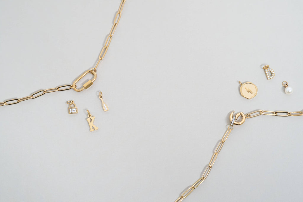 Two gold chain necklaces by WJW, including the sleek 18K Gold PVD Paperclip Carabiner Necklace, are arranged on a light surface with several small gold and pearl charms nearby—featuring designs like a key, lock, letter K, and round pendant. Perfect for customizing your look or layering with your favorite Carabiner Charm Holder Necklace for added style.
