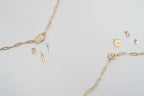 Two gold chain necklaces by WJW, including the sleek 18K Gold PVD Paperclip Carabiner Necklace, are arranged on a light surface with several small gold and pearl charms nearby—featuring designs like a key, lock, letter K, and round pendant. Perfect for customizing your look or layering with your favorite Carabiner Charm Holder Necklace for added style.