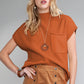 A woman with long blonde hair wears Shewin's Patch Pocket Ribbed Knit Short Sleeve Sweater in orange and a tan skirt, accessorized with a wooden necklace and bracelet. She stands against a neutral background, perfect for transitional weather.