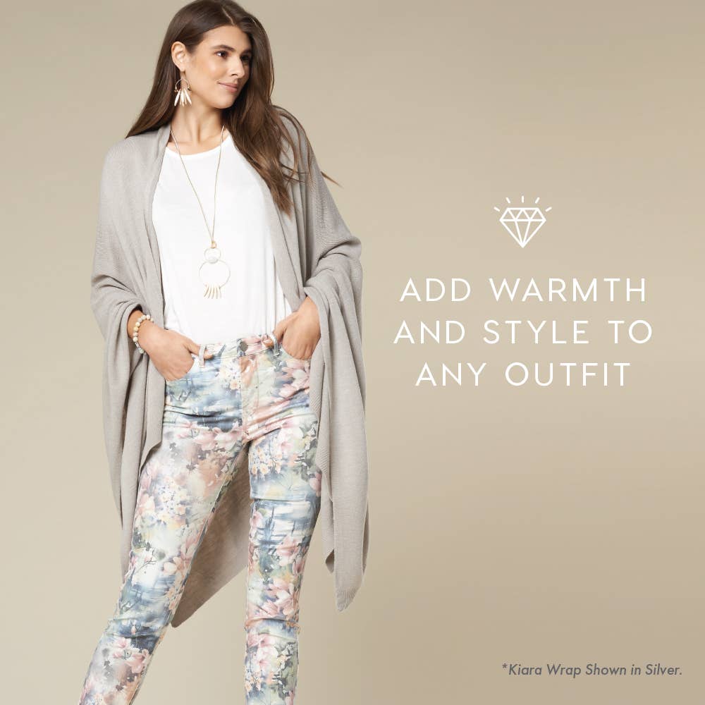 A woman in a COCO + CARMEN Kiara Light Weight Wrap in silver, layered over a white top and floral pants, stands before a beige backdrop. Text: "Add warmth and style to any outfit. *Kiara Light Weight Wrap in Silver, one size fits most.