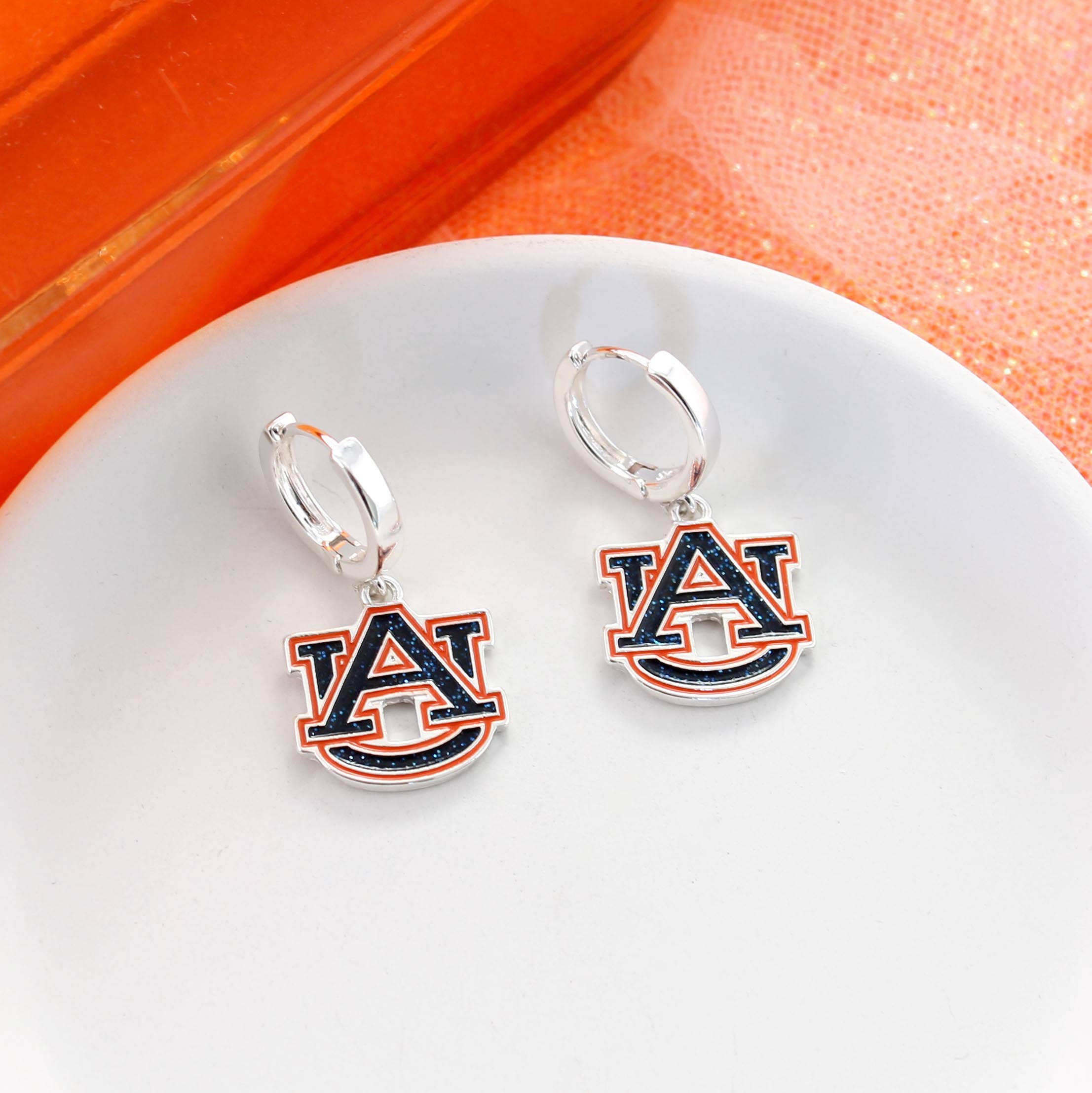 A pair of Auburn Glitter Enamel Logo Huggie Hoop Earrings by Seasons Jewelry, featuring the Auburn University logo in a sparkling silver huggie hoop design—perfect game day jewelry for any fan, displayed on a white plate with an orange background.