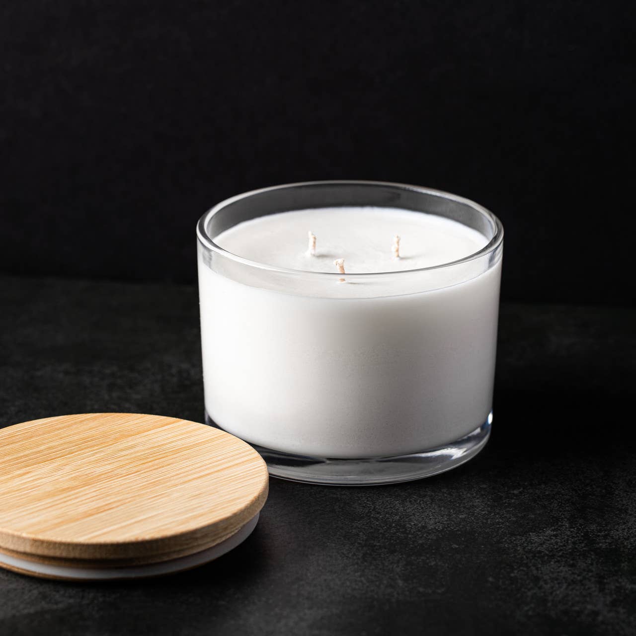 A white 20oz three-wick soy wax candle in a clear glass jar from Stephanie Henry Candle Company rests on a dark surface, offering a Coconut Lime scent. The wooden lid is placed beside it.