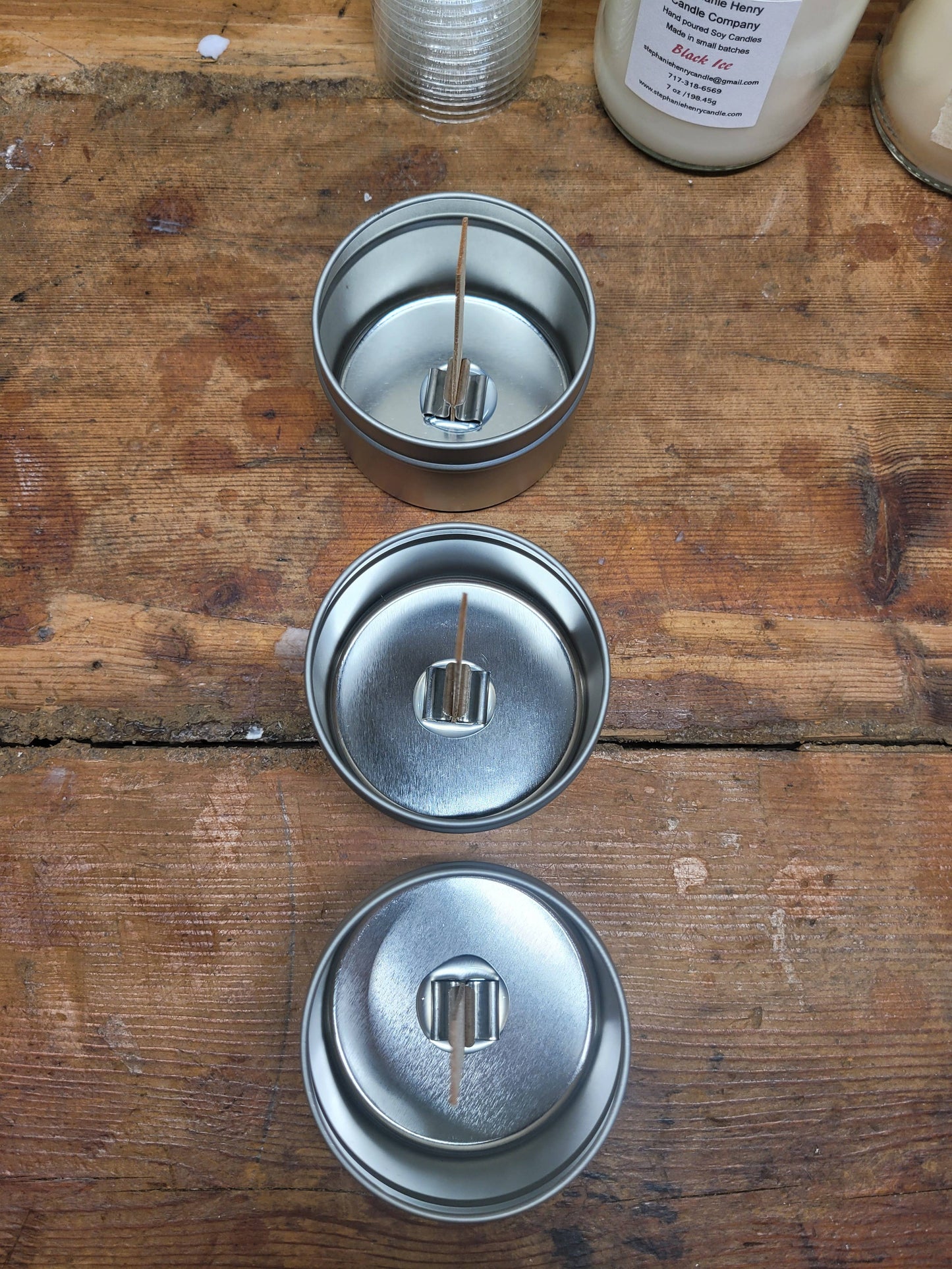 Three 4oz Handpoured Soy Wax Candles by Stephanie Henry Candle Company, in metal tins with wood wicks, rest elegantly on a wooden surface when viewed from above.