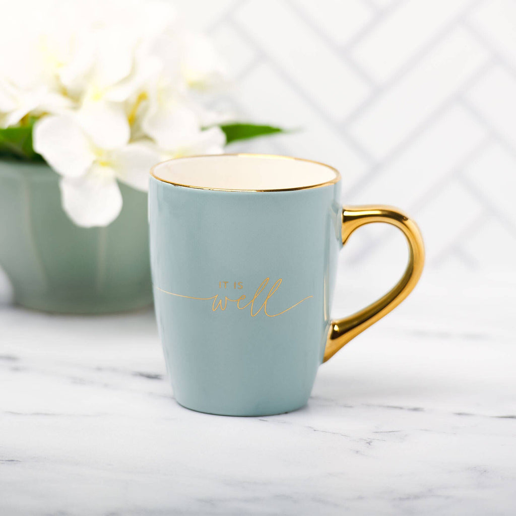 A beautiful Mug Blue/Cream "It is Well with My Soul" from Christian Art Gifts sits on a white marble surface, featuring elegant gold accents and a gold handle. The inspiring message “It is well with my soul” appears in gold text on the light blue and cream ceramic mug, while a vase of white flowers provides a lovely background touch. This charming mug is perfect for anyone who cherishes this classic faith-filled reminder.