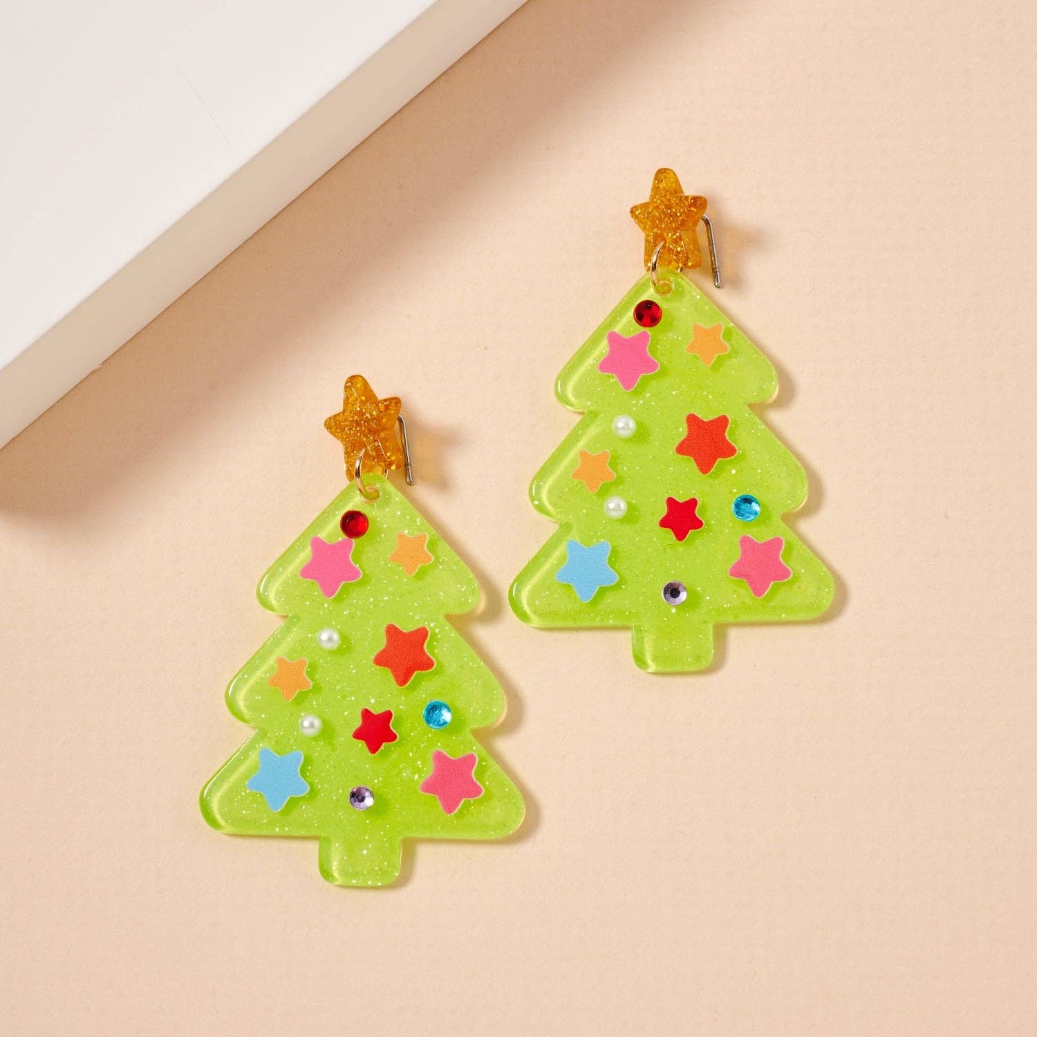 The Glittery Christmas Tree Drop Earrings from Lilla Haven are festive holiday statement pieces, crafted from glimmering green acrylic in the shape of Christmas trees and adorned with colorful stars, vibrant gems, and orange star toppers. These eye-catching earrings are beautifully displayed on a light beige surface. Main image